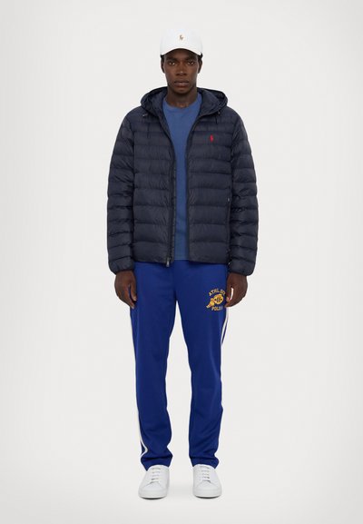 Giacca puffer blu navy con cappuccio, t-shirt blu sotto, pantaloni da jogging blu con logo giallo, e sneakers bianche. Design semplice e lineare.