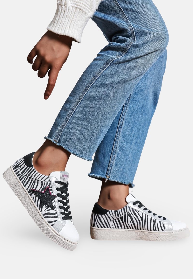 Baskets à motif zèbre avec un bout en cuir blanc, des côtés noirs, un détail en étoile rose et une semelle en caoutchouc texturé, portées avec un jean bleu.