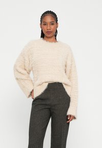 Maglione fluffy beige chiaro con colletto rotondo, vestibilità morbida e polsini a coste, abbinato a pantaloni high-waisted scuri e strutturati.