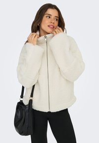 Kvinna som justerar kragen på en vit fleece-zip-up jacka, iklädd svarta byxor och bär en svart axelväska.