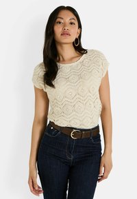 Top de encaje crema con un patrón de olas, mangas cortas y escote redondeado, combinado con jeans azul oscuro y un cinturón marrón con una hebilla circular.