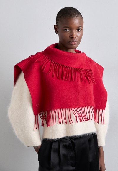 Poncho en mélange de laine rouge avec des détails à franges à l'ourlet, associé à un haut blanc doux et texturé et un pantalon noir plissé.