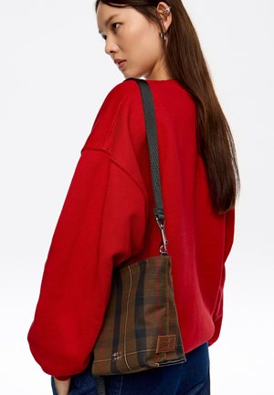 Bolso de cuadros marrón con forma estructurada, que cuenta con una larga correa negra y una etiqueta de acento de cuero. El modelo lleva una sudadera roja.