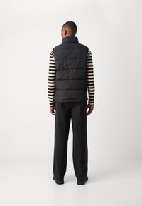 Black, quilted puffer vest with high collar, worn over a striped long-sleeve shirt, paired with wide-leg black pants, viewed from the back.