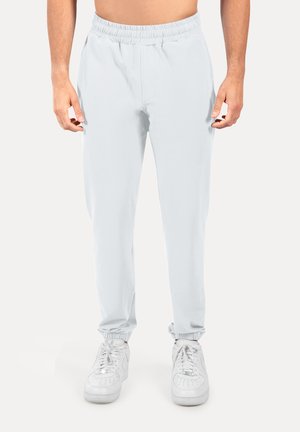 Mann trägt weiße Jogginghose mit elastischem Bund und weiße Sneakers, steht vor einem schlichten Hintergrund.