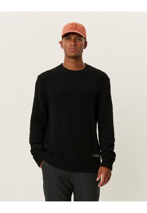 Schwarzer strukturierter Strickpullover mit Rundhalsausschnitt und gerippten Bündchen, mit einem dezenten gestickten Logo am unteren Rand. Dazu eine Mütze.