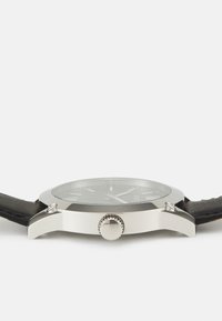 Silberner Edelstahl-Uhren mit einem runden schwarzen Zifferblatt, leuchtenden Stundenmarkierungen und einem strukturierten schwarzen Lederarmband. Enthält eine geriffelte Krone.