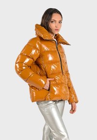Veste matelassée orange brillante avec un col haut et une fermeture éclair, présentant une texture matelassée et des manches ajustées. Un pantalon argent complète le look.