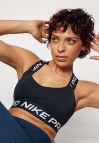 Crna sportska grudnjak s izrezom u obliku suze, kontrastnim bijelim "PRO" i "NIKE" oznakama na trakama i pojasu, glatke teksture.
