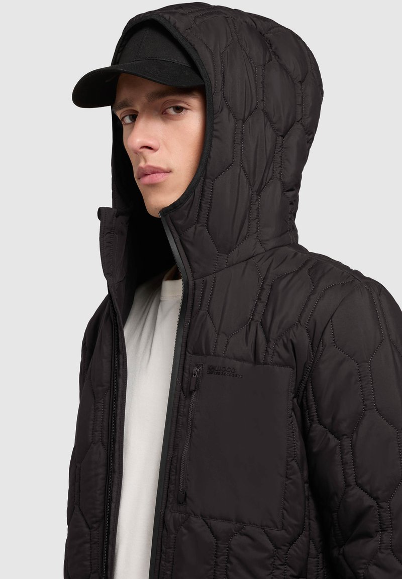 Veste à capuche noire matelassée avec motif hexagonal, fermeture éclair à l'avant et poche poitrine zippée. Texture douce, conçue pour la chaleur.