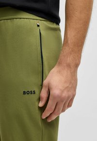 Olijfgroene broek met een zachte textuur, zijzak met rits en geborduurd "BOSS" logo in donkerblauw nabij de zak.