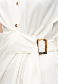 Κοντινό πλάνο λευκού φορέματος με ζώνη με αγκράφα από χελώνιο και καφέ κουμπιά μπροστά.