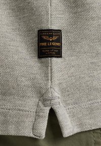 Tejido de punto gris claro con una pequeña etiqueta negra y dorada que dice "PME Legend, Probado a Gran Altura". Presenta un diseño con abertura lateral.