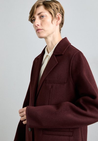 Cappotto di lana bordeaux con colletto a punte, tasca sul petto singola e spalle scese. Presenta una texture liscia e chiusura con bottoni.