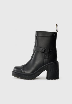 Botas de tornozelo em couro preto com um salto alto robusto, detalhe de fivela lateral e tachas decorativas ao longo da costura frontal. Sola de borracha texturizada.