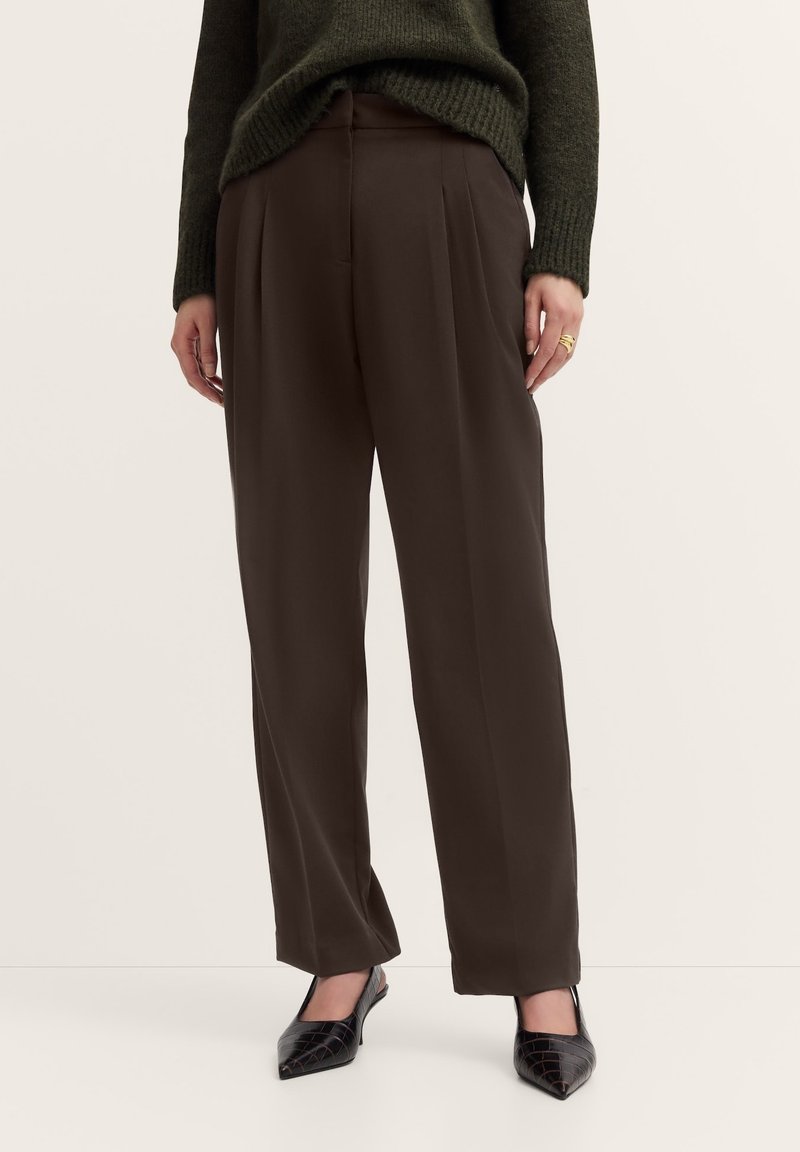Brown pleated trousers with a high waist, made from smooth fabric, paired with black pointed flats and a knitted dark green sweater.