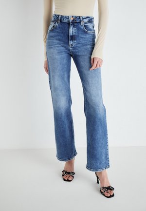 Kvinde iført blå højttaljede jeans med lige ben kombineret med sorte høje hælede sandaler med perlebesatte sløjfer.