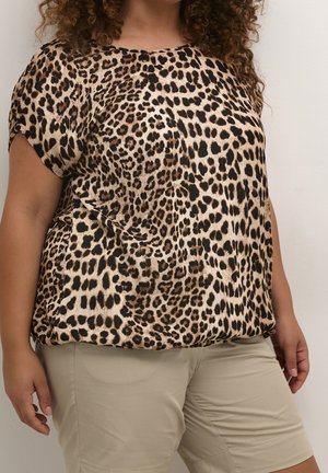 Mujer con una blusa de manga corta con estampado de leopardo y pantalones beige hasta la rodilla, de pie frente a un fondo liso de color claro.