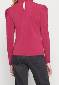 Long-sleeve top in fuchsia, featuring gathered shoulder detailing, a high collar, and a back keyhole opening. Smooth, stretchy fabric.