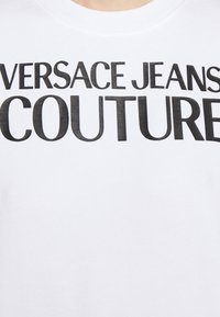 Bílá mikina s výrazným černým textem s nápisem "VERSACE JEANS COUTURE." Materiál vypadá měkce a má žebrovaný výstřih.