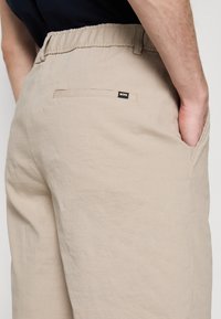 Beige puuvillashortsit joustavalla vyötäröllä, sivutaskuilla ja pienellä mustalla logopatchilla. Yksinkertainen muotoilu, jolla on sileä pinta.