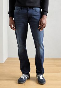 Tummansiniset farkut slim fit -mallilla, joissa on lievää kulumista, viisi taskua ja ommeltuja yksityiskohtia. Yhdistetty tummiin tennareihin, joissa on valkoiset raidat.