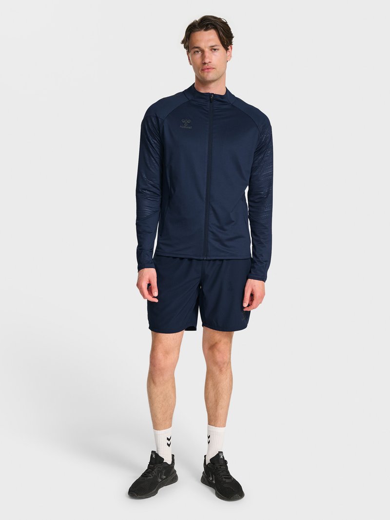 Homme debout portant une veste de sport bleu marine zippée, un short assorti, des chaussettes blanches et des chaussures de sport noires sur fond uni.
