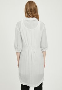 Rochie albă, lejeră, cu guler, mâneci trei sferturi și talie elastică. Textură ușoară, care ajunge sub genunchi.