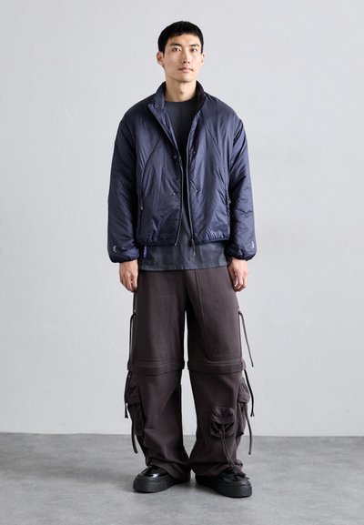 Giacca puffer blu navy con colletto alto, chiusura a zip e orli elasticizzati, abbinata a pantaloni cargo marroni larghi con tasche e cordini.