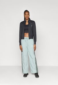 Juodas zip-up džemperis, juodas trumpas topas, šviesiai mėlynos kelnės su tamsiai žaliais šoninėmis detalėmis ir juodi batai. Sportiškos medžiagos ir laisvas kirpimas.