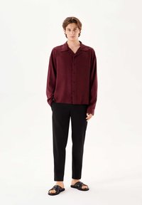 Cămașă burgundy cu butoni, cu guler, mâneci lungi și o croială lejeră, asortată cu pantaloni negri slim-fit și sandale negre.