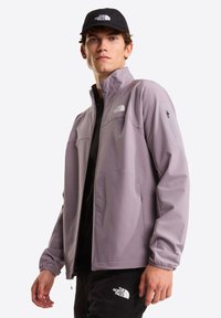 Giacca grigio chiaro con zip frontale, due tasche laterali, texture a diamante sottile e logo sul petto. Abbinata a una maglia nera e un cappellino.