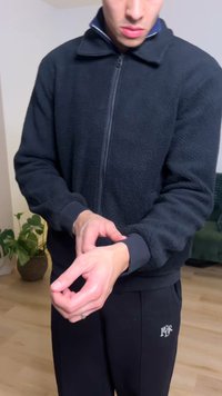 Veste polaire noire à fermeture éclair avec poignets côtelés, portée sur un pantalon de survêtement noir avec un logo. Mains placées près de la zone des poignets.