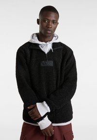 Sudadera negra de forro polar con cuello alto, media cremallera y parche con el logo de Vans. Capa con una sudadera gris debajo, que presenta puños acanalados.