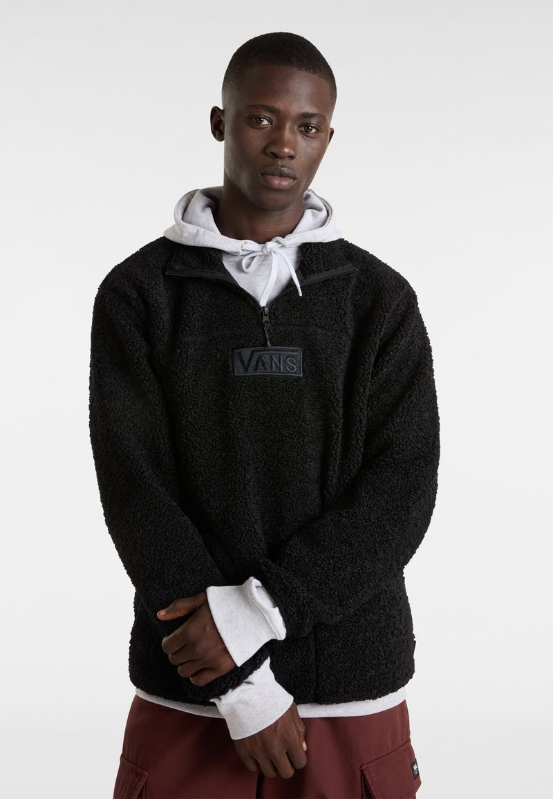 Sudadera negra de forro polar con cuello alto, media cremallera y parche con el logo de Vans. Capa con una sudadera gris debajo, que presenta puños acanalados.