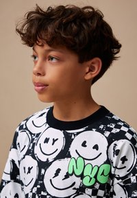Chemise à manches longues noir et blanc arborant un motif de visage souriant et un logo "NYC" vert, confectionnée en coton doux.