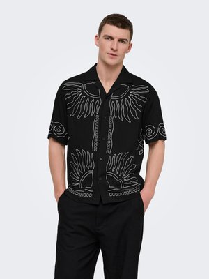 Jonge man draagt zwart kortemouwshirt met witte abstracte zon- en spiraalpatronen, staat met handen in de zakken, neutrale achtergrond.