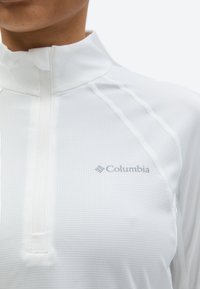 Persona, kas valkā baltu Columbia garu piedurkņu apģērbu ar ceturtdaļas rāvējslēdzēju un zīmola logo uz krūtīm, ar teksturētu audumu un šuvju detaļām.