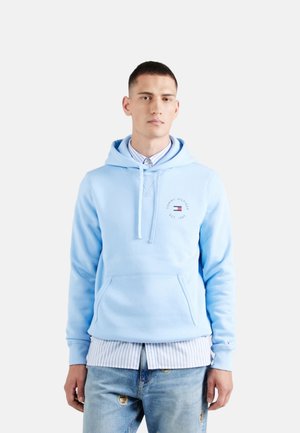 Sudadera azul claro fabricada en una mezcla de algodón, con un bolsillo frontal, capucha con cordón y un acento de logotipo. Combinada con una camiseta de rayas por debajo.