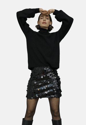 Black knitted sweater with a high neck and long sleeves paired with a sequined black mini skirt featuring circular reflective accents.