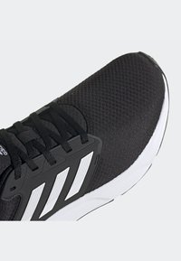Scarpa da ginnastica nera con tomaia in mesh testurizzato, caratterizzata da audaci accenti bianchi, lacci rotondi e una suola bianca ammortizzata.