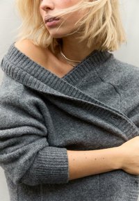 Siv pulover iz volne z rebrastim off-shoulder dizajnom in zloženim ovratnikom. Tekstura se zdi mehka, z oprijetimi rokavi in sproščenim krojem.