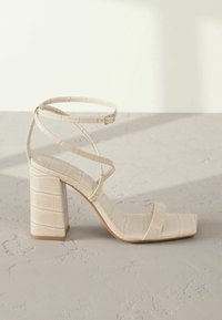Sandale à talon haut beige avec un bout carré, un motif texturé effet crocodile, un talon carré épais, et des brides croisées avec une boucle réglable.