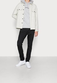 Camicia overshirt in flanella grigio chiaro con due tasche sul petto, indossata sopra un hoodie grigio, abbinata a jeans skinny neri e sneakers bianche.