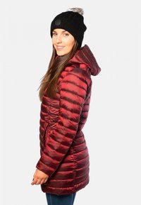 Veste matelassée rouge avec une texture brillante, dotée d'une capuche et de poches latérales, associée à un bonnet en tricot noir avec un pompon.