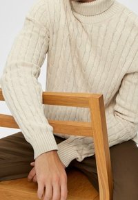 Lichtbeige kabelgebreide coltrui met geribde manchetten, gedragen door een zittende figuur die zijn hand op een houten stoel legt.