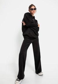 Schwarzer übergroßer Hoodie mit Fronttasche, kombiniert mit schwarzen Schlaghosen. Das Model trägt schwarze Sonnenbrillen und weiße Sneakers mit schwarzen Akzenten.