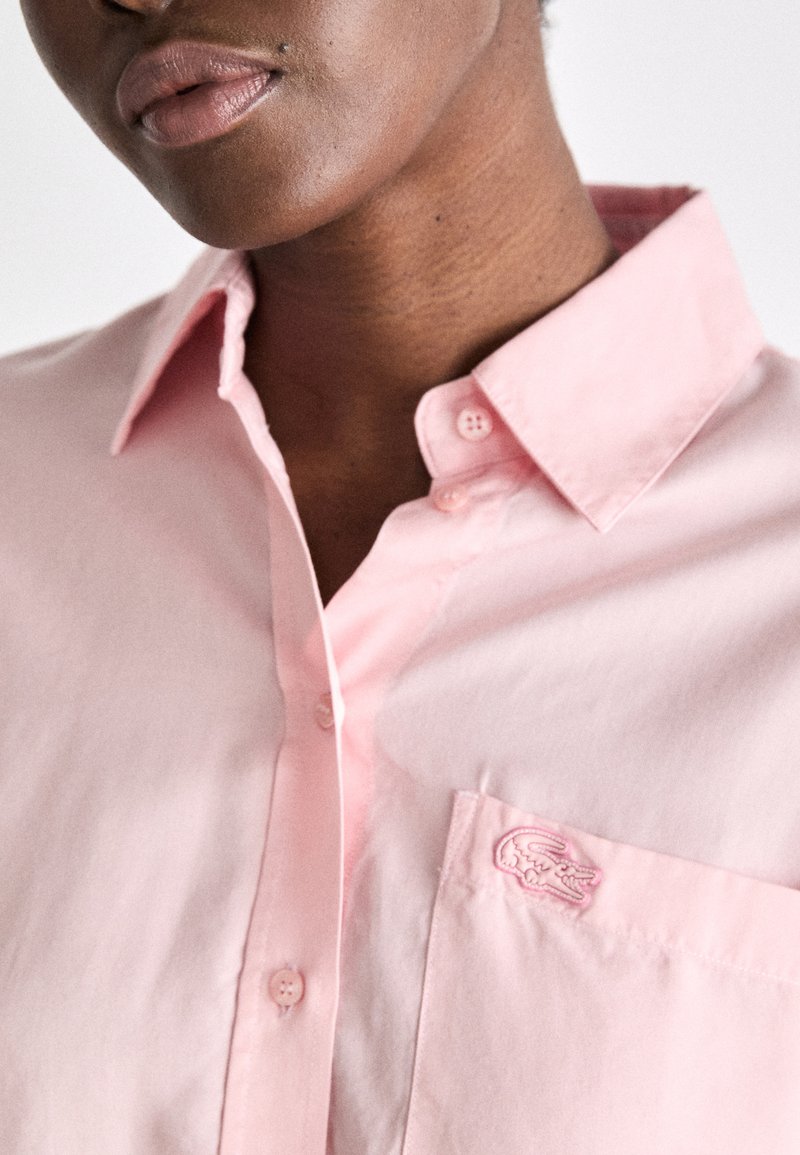 Chemise boutonnée rose clair avec un col, présentant une poche avec un logo brodé. Le tissu semble lisse et légèrement texturé.