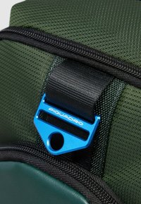 Detalle de bolsa de tela verde con un clip metálico azul, cremallera negra y superficie texturizada; presenta bordes cosidos y una sección de acento lisa.