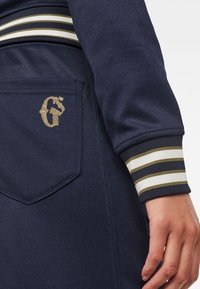 Mörkblå sweatshirt med guldbroderad logotyp på fickan, med ribbade ärmslut med vita och olivgröna ränder för extra detalj.